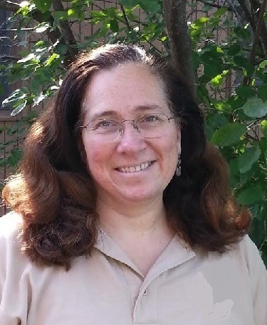 Smiling woman with long, wavy hair, wearing glasses and a light-colored shirt, stands outdoors among green foliage.