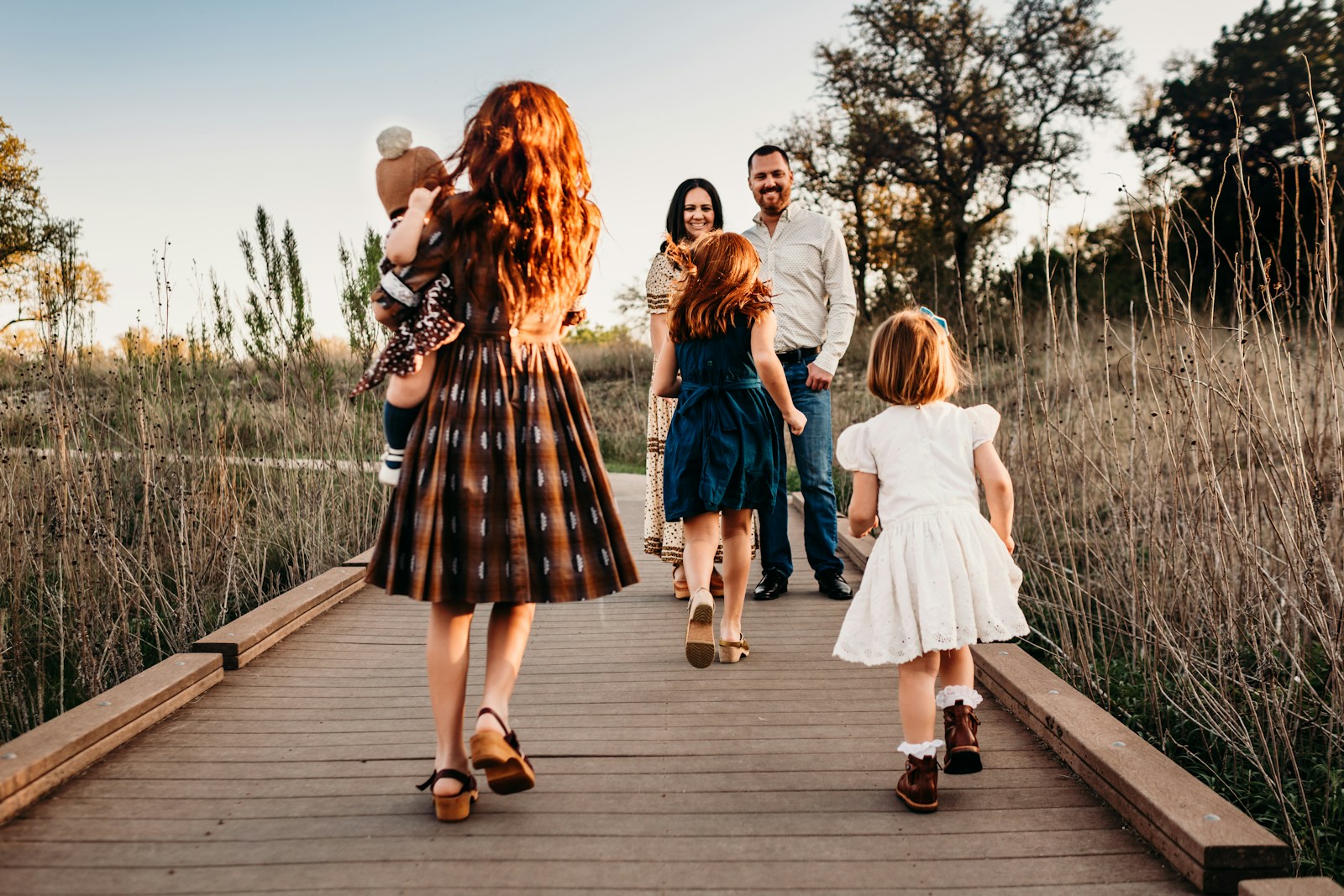 A family walks together on a wooden path, surrounded by tall grass and trees, capturing a moment of togetherness during golden hour.
