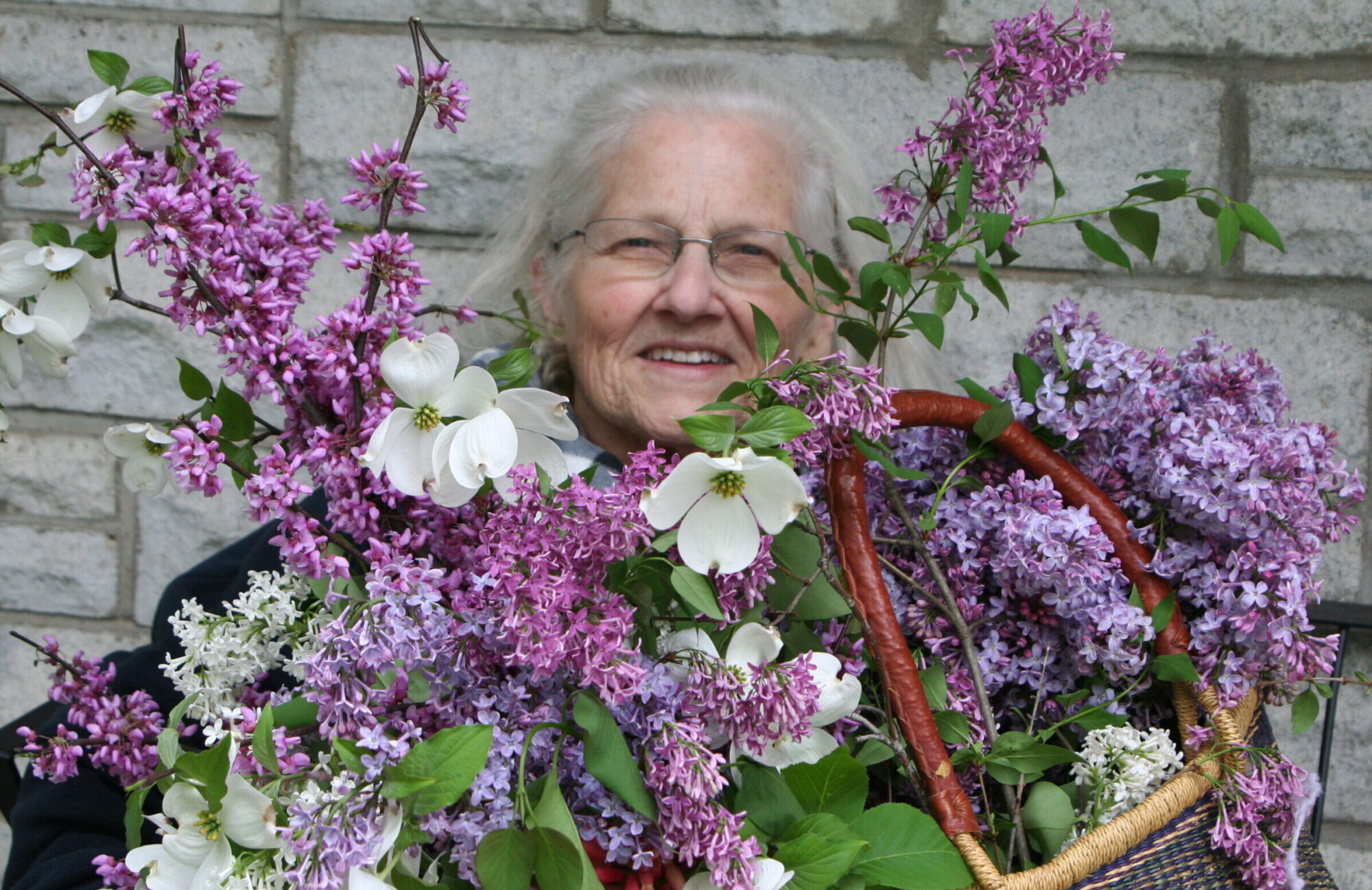 Elderly woman smiling, surrounded by vibrant purple lilacs and white dogwood flowers, showcasing a beautiful spring arrangement.