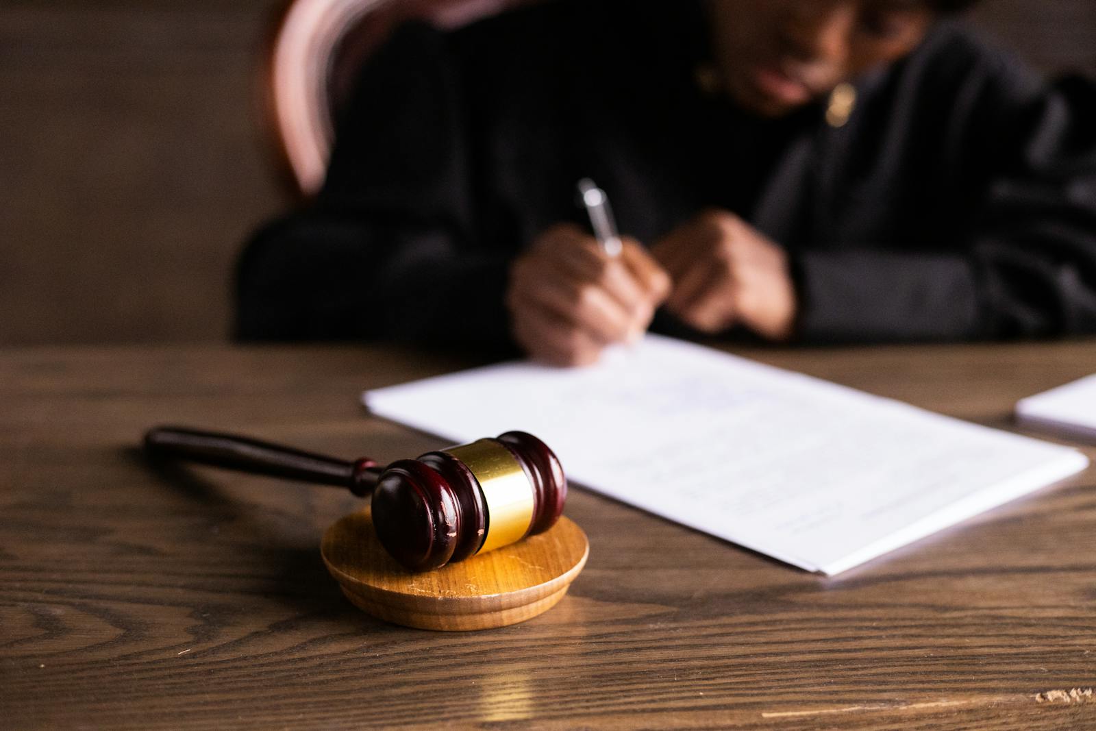 A gavel rests on a wooden block beside a person in a black robe, who is writing on documents. The image represents legal authority and proceedings.